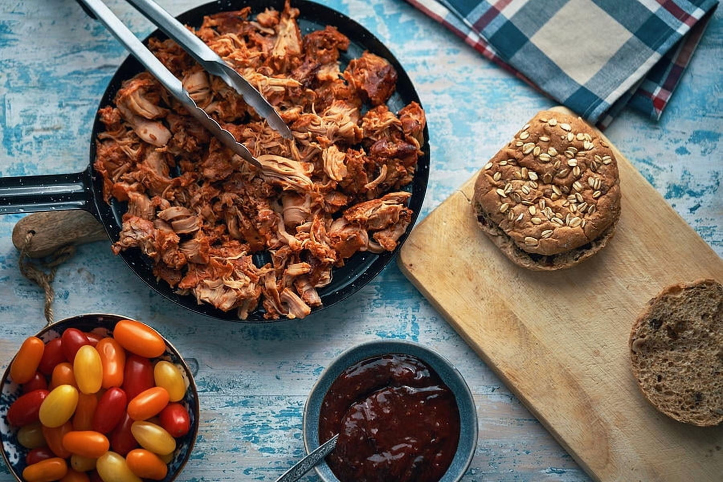 Shredded pulled pork in a black skillet with metal tongs, served on a rustic blue table with a sandwich on a wooden board, a bowl of cherry tomatoes, and a bowl of sauce.