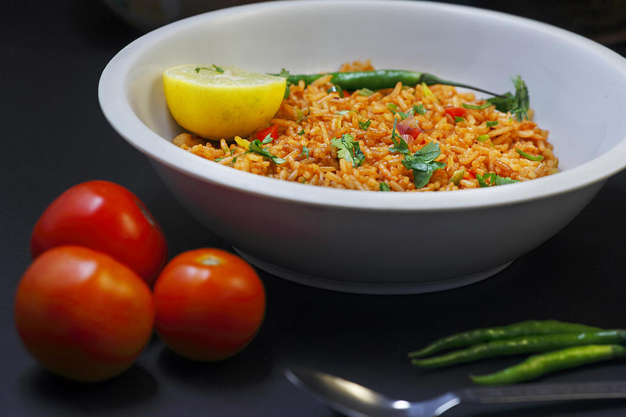 A white bowl filled with Portuguese Tomato Rice, garnished with green herbs, red peppers, and a lemon wedge, set on a dark surface with tomatoes, green chilies, and a spoon nearby.