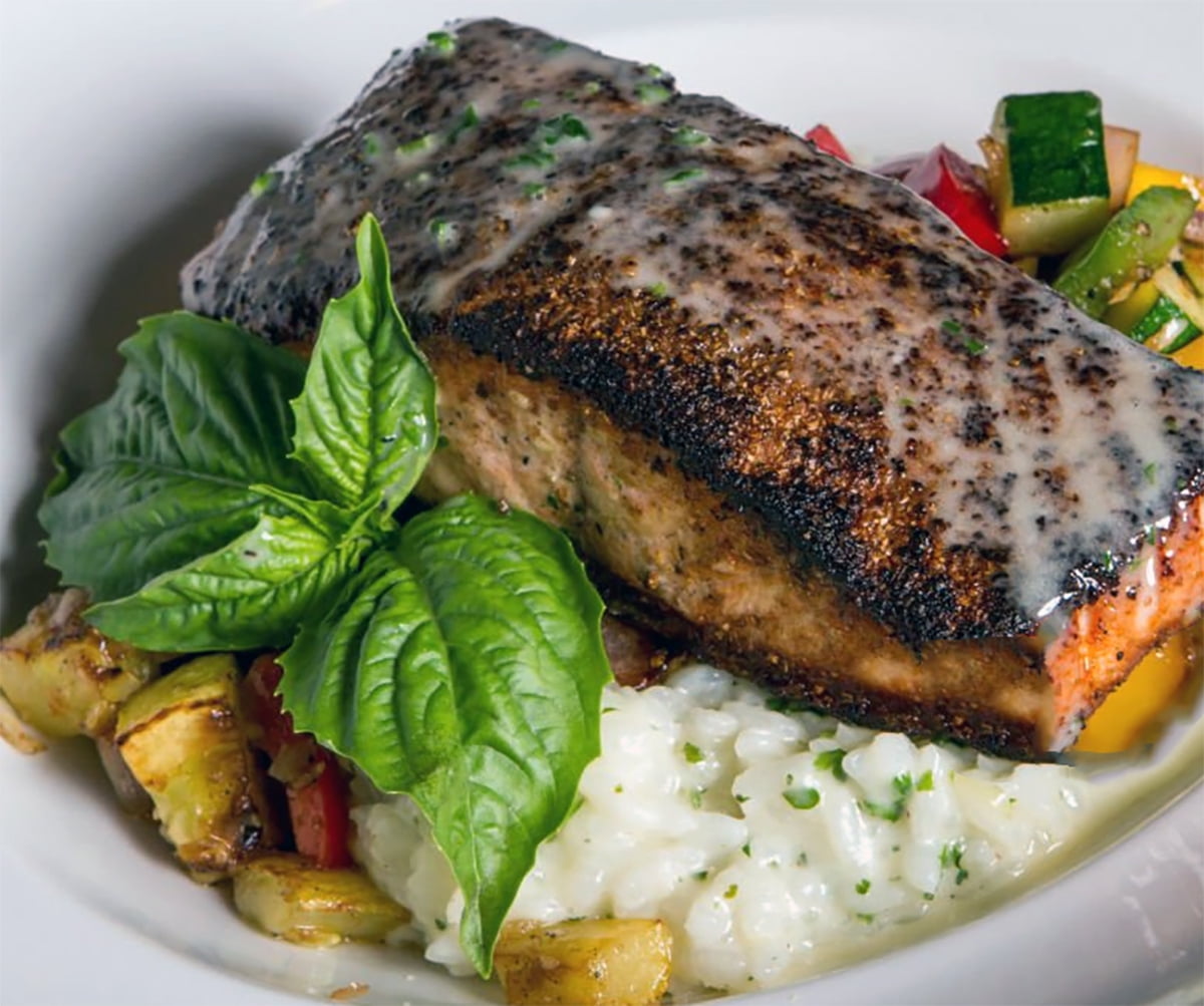 A plated salmon fillet with a charred blackened crust, topped with a creamy sauce, served over white risotto with mixed vegetables, green beans, and fresh basil leaves.