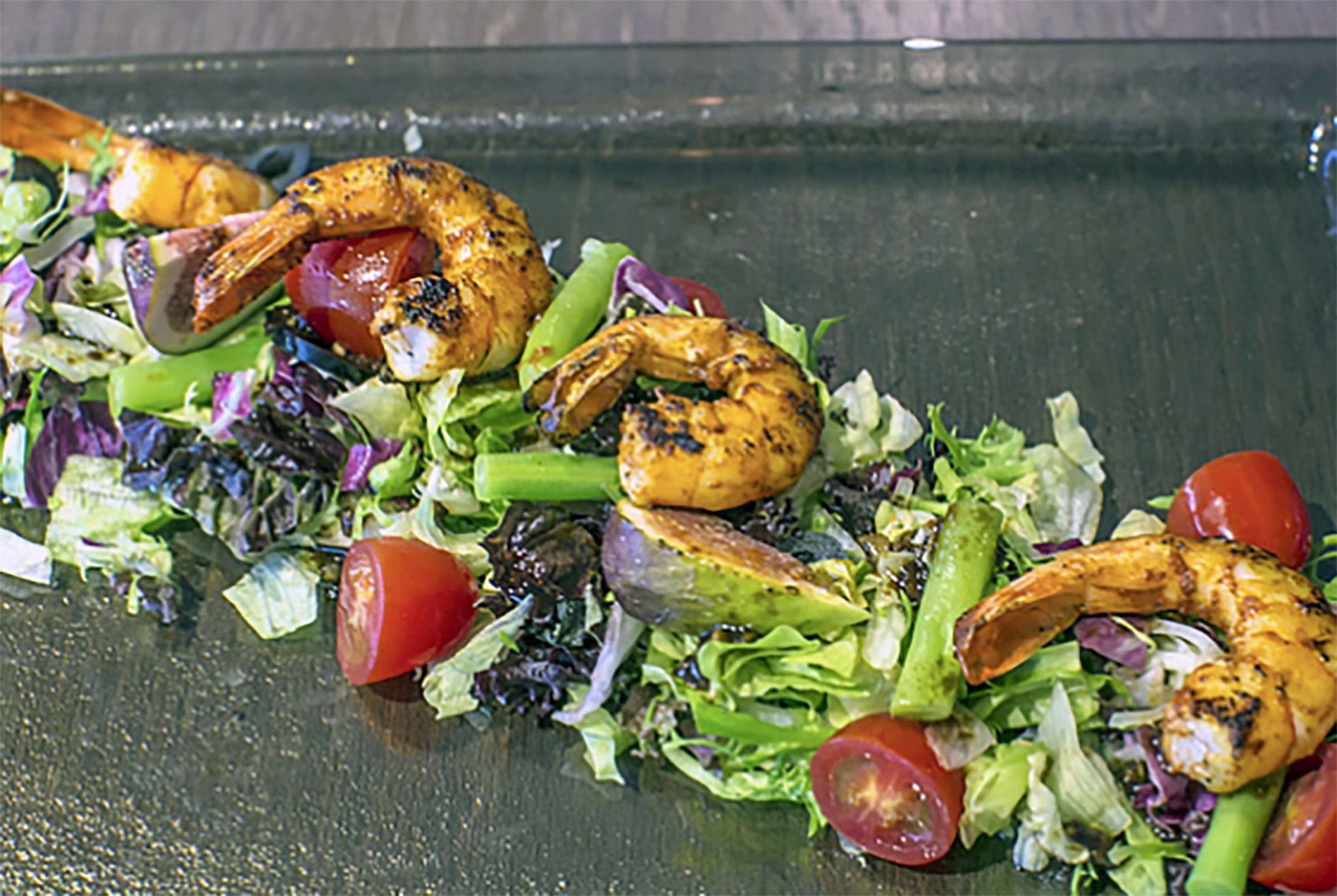 Grilled shrimp salad on a dark slate platter, featuring cooked shrimp, mixed greens, cherry tomatoes, and asparagus.