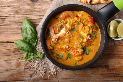 A top-down view of a black frying pan filled with Authentic Portuguese Shrimp Mozambique served with crusty bread and buttery garlic sauce, set on a rustic wooden table with basil leaves and lime wedges.